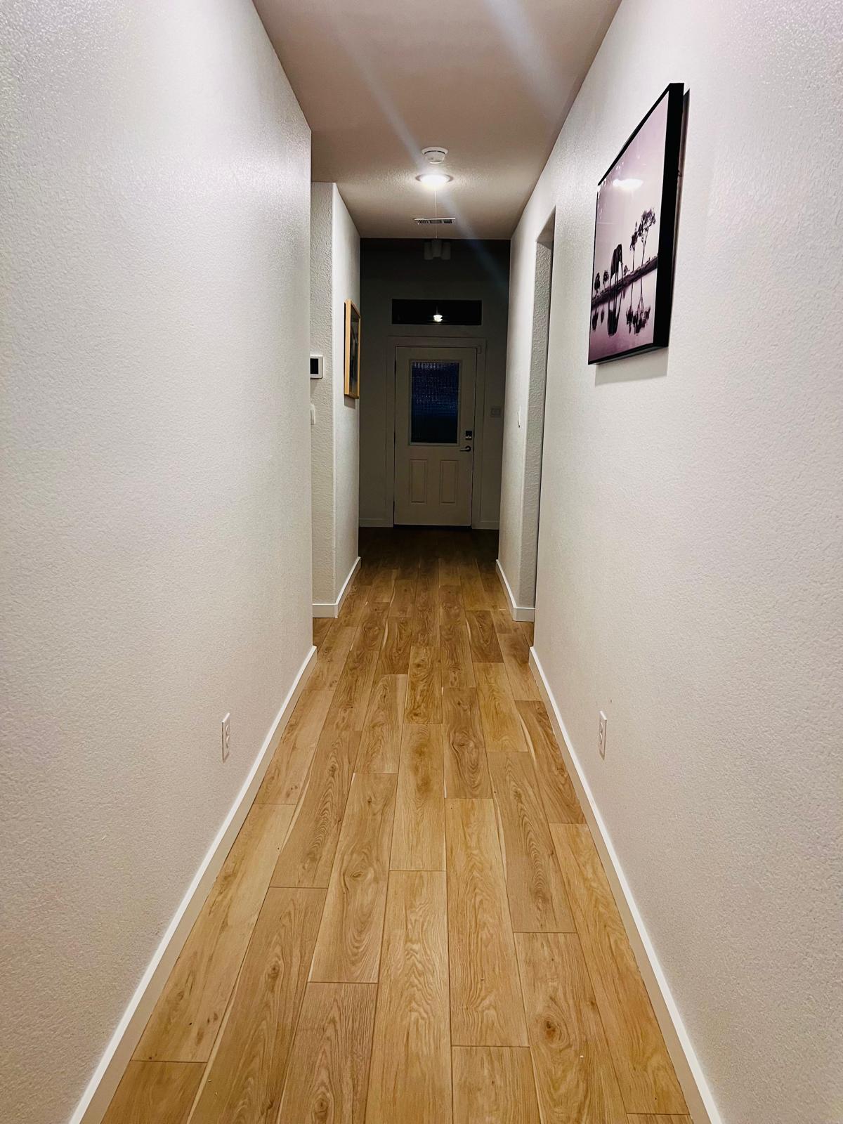 Bright hallway with light wood-look flooring, white walls and framed artwork leading to a door